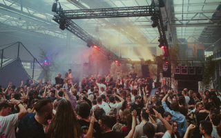 Crowd of people dancing at an indoor concert with colorful stage lights and smoke effects.