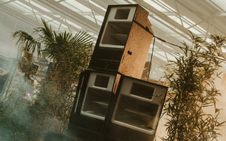 Large speakers surrounded by lush green plants in a greenhouse setting.